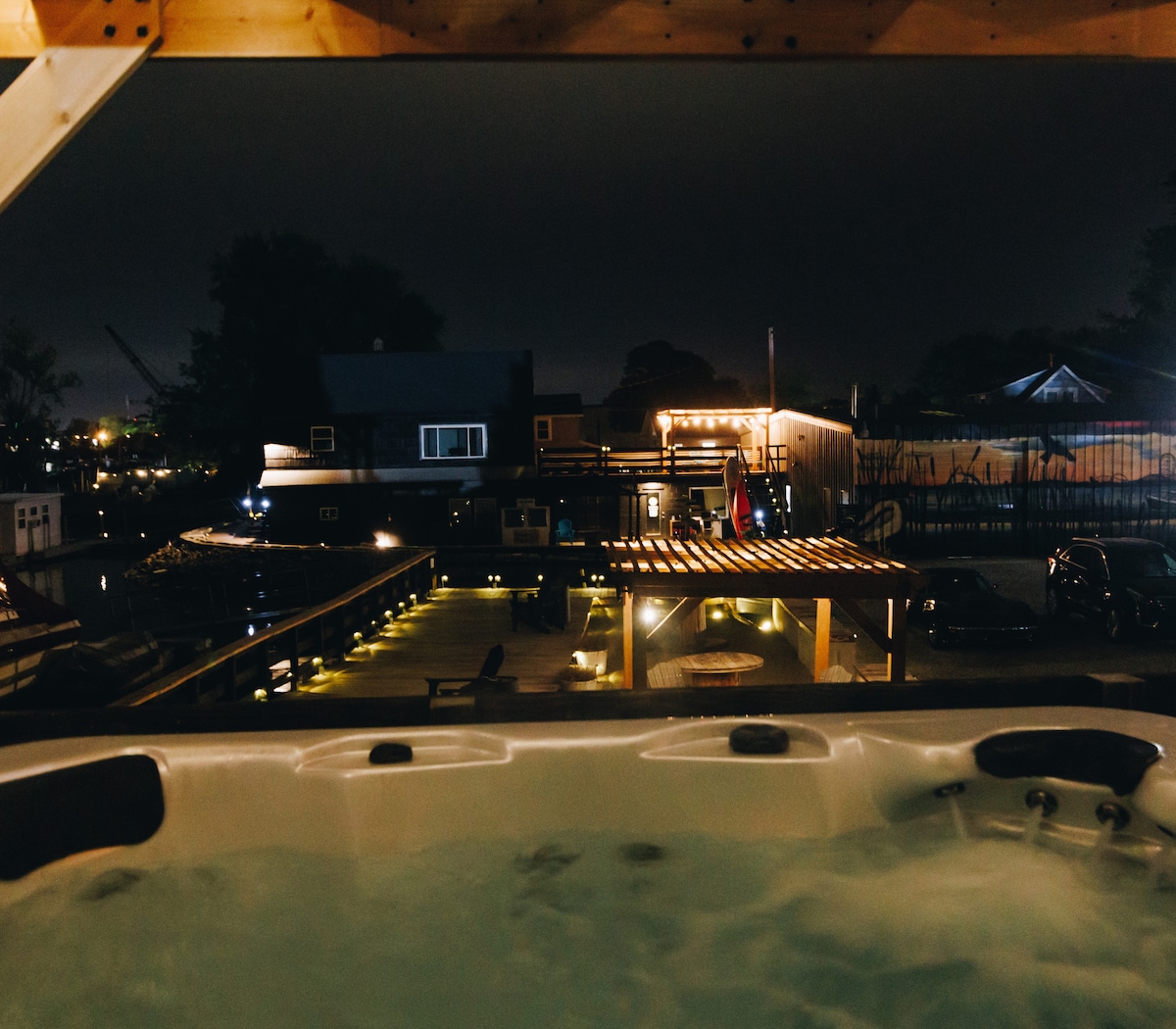 The image showcases a private hot tub with water visible, surrounded by wooden decking. Soft lighting illuminates the nearby boats and structures in the marina, creating a serene evening atmosphere. The view highlights a tranquil retreat experience amidst the gentle movement of water.