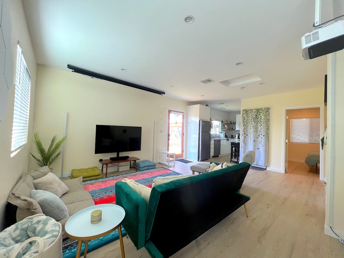 A spacious living area features a comfortable sofa and a vibrant area rug, creating a relaxed atmosphere. A television is positioned against the wall, while a doorway leads to the kitchen area, which is visible in the background. Natural light enters through the windows.