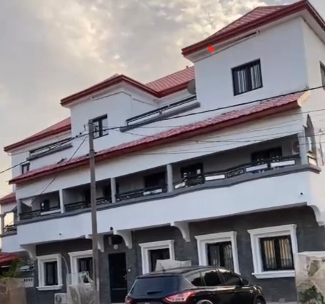 The exterior of a three-story building is presented, showcasing a combination of white and grey walls. The structure features a red-tiled roof and multiple windows with black frames. A vehicle is parked in front, while the surrounding area includes a sidewalk and fencing.