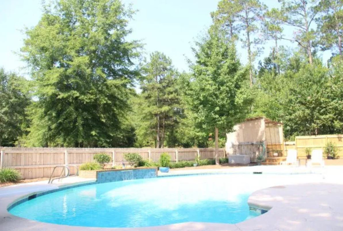A serene outdoor area is showcased with a clear gunite saltwater pool, surrounded by lush greenery and mature trees. A spacious patio area offers ample space for relaxation, complemented by a pergola in the background, and a wooden fence encloses the space for privacy.