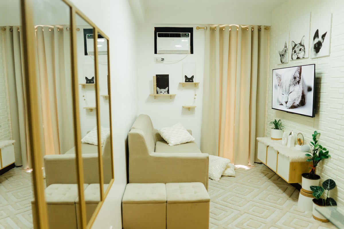 A cozy living area features a light-colored sectional sofa adorned with soft pillows. A flat-screen TV mounted on a white brick wall displays a feline image. Two floating shelves display decorative items, complemented by houseplants beside a mirrored cabinet, enhancing the room's brightness.