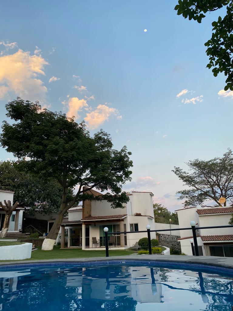 The image captures a serene outdoor scene featuring a clear blue pool. In the background, a two-story house can be seen along with lush green trees and a soft sky illuminated by the moon and clouds at dusk.