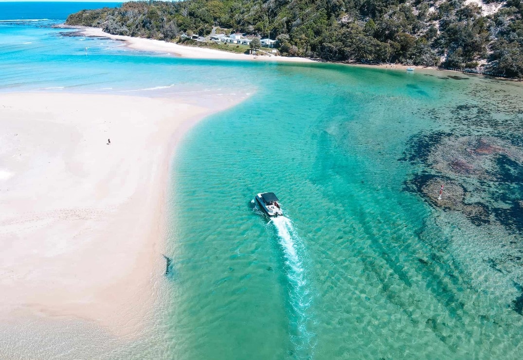 A tranquil waterway glimmers under the sun, revealing soft sandy banks that curve into the vibrant turquoise sea. A boat glides smoothly through the clear water, surrounded by natural beauty and lush greenery in the distance.