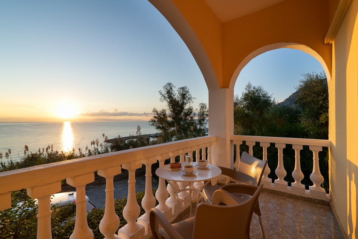 A private balcony offers scenic views of the sea during sunset, framed by arched columns. A small round table is set with two chairs, inviting relaxation. Lush greenery is visible in the background, enhancing the calm atmosphere of this coastal retreat.