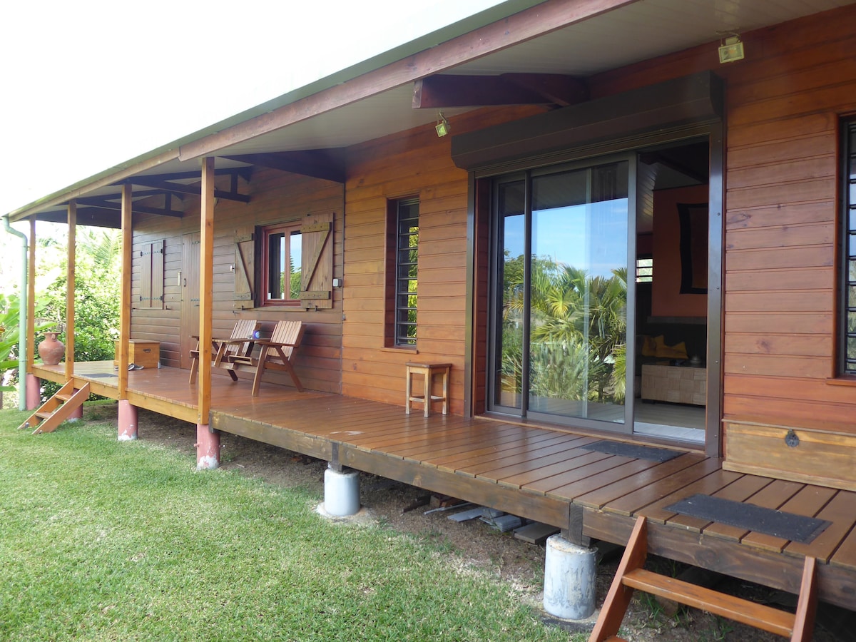 A wooden building features a spacious covered terrace with a wooden deck. Two chairs and a small table are placed on the patio, with sliding glass doors providing access to the interior. Lush greenery surrounds the area, enhancing the natural setting.