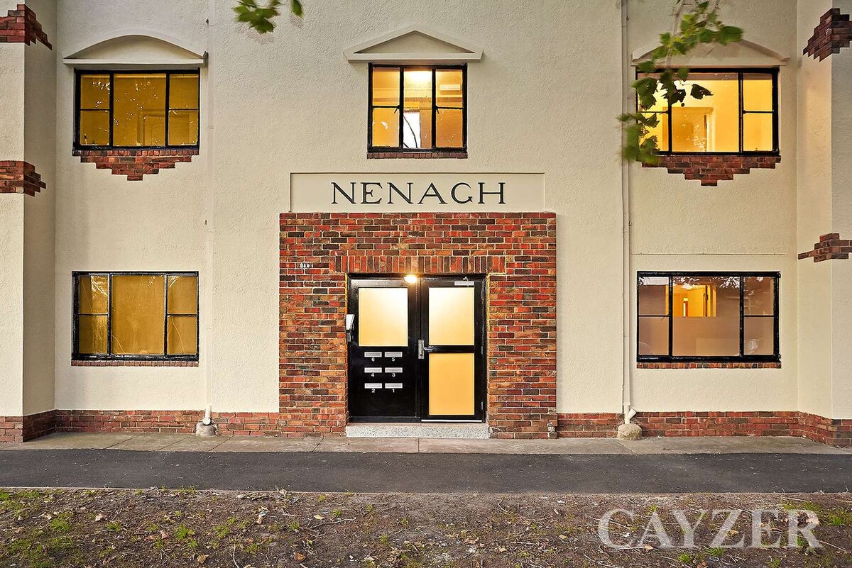 The exterior of a two-story building is shown, featuring a combination of brick and cream-colored walls. The entrance is marked with the name 'Nenagh,' and large windows are visible on both levels, framed in dark trim. Surrounding greenery adds to the setting.