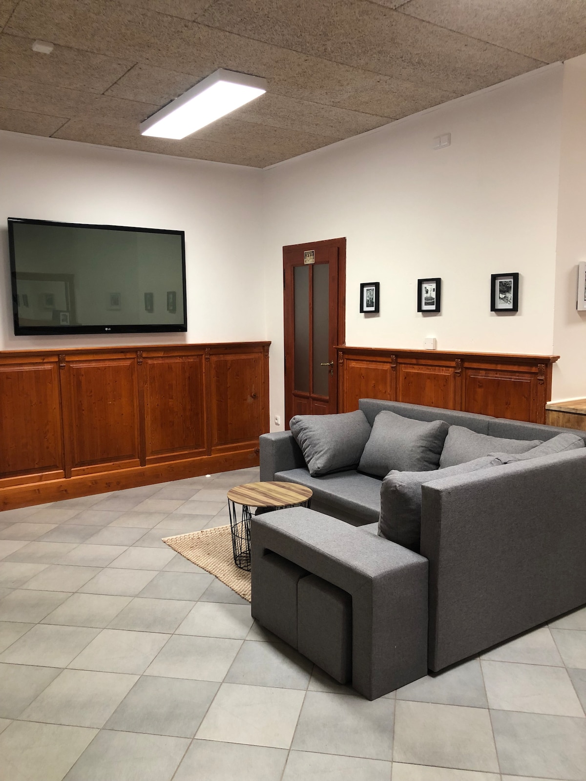 A cozy seating area features a gray sofa accompanied by a small round table. A wall-mounted television is positioned above a wooden panel, providing a touch of modernity. Framed photos are displayed on the adjacent wall, while the floor is covered with neutral-toned tiles.