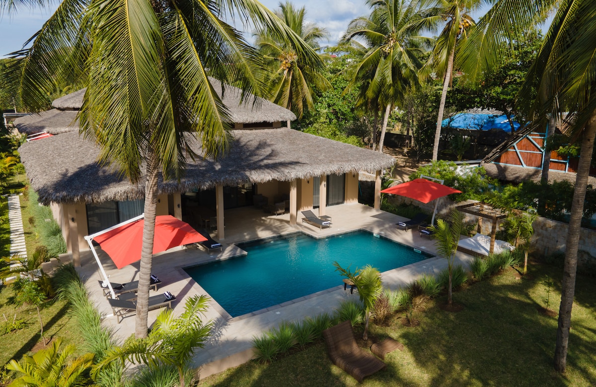 A modern villa features a thatched roof and is surrounded by lush greenery. A rectangular pool invites relaxation, with sun loungers positioned nearby. Umbrellas provide shade while palm trees sway gently in the breeze, creating an inviting outdoor space.