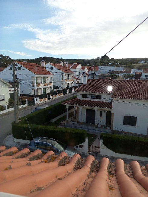 A view from a rooftop showcases neighboring homes with red-tiled roofs and well-maintained gardens. Street scenes feature a car parked on a quiet road. A clear sky with wispy clouds enhances the tranquil atmosphere of this residential area.