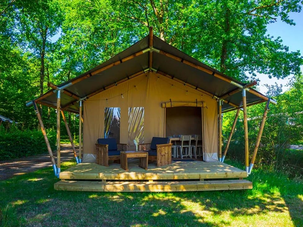 A spacious tent structure is supported by wooden beams, featuring a covered porch area. Two chairs are positioned around a small table on the wooden deck, while large windows allow natural light to enter. Surrounding greenery enhances the tranquil setting.