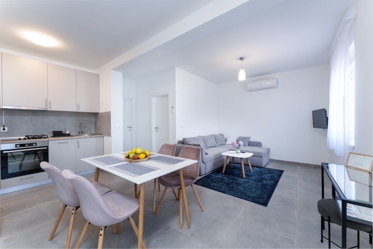 A modern living space features a light-filled environment with a grey sofa arranged around a coffee table. A dining area with a white table and four chairs is present, enhancing the inviting atmosphere. The kitchen, visible in the background, includes a stove and cabinetry.
