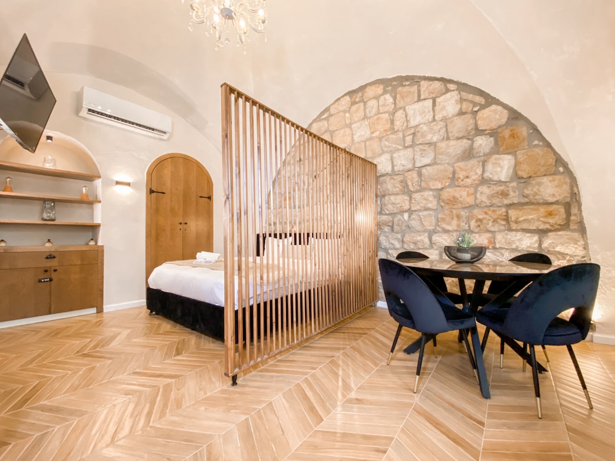 An open-layout studio showcases Jerusalem stone walls beneath an arched ceiling. A wooden screen provides subtle separation between the sleeping area with a queen bed and a dining space with a round table and four chairs. Natural light brightens the warm wooden floor.