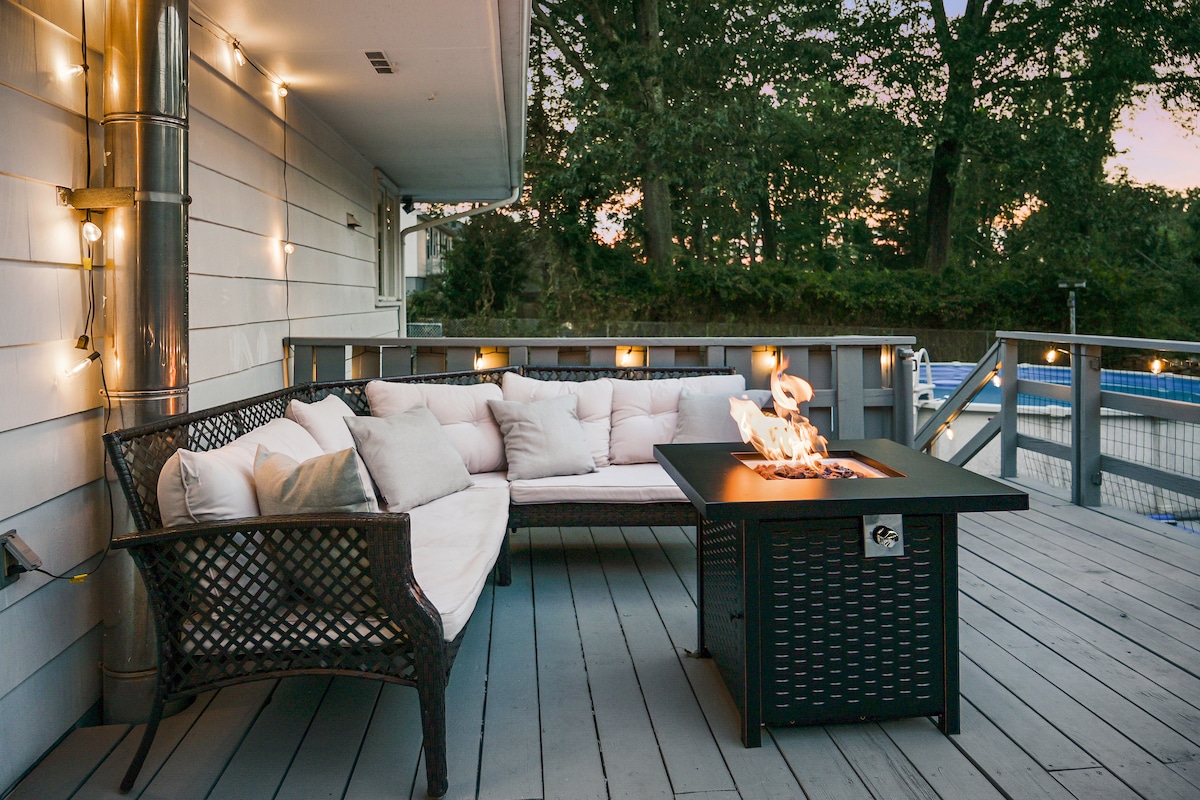 An outdoor seating area features a comfortable sectional sofa adorned with soft, white cushions. A fire pit sits at the center, with flames dancing atop a sleek black surface. Soft string lights provide a warm glow, creating an inviting atmosphere for relaxation.