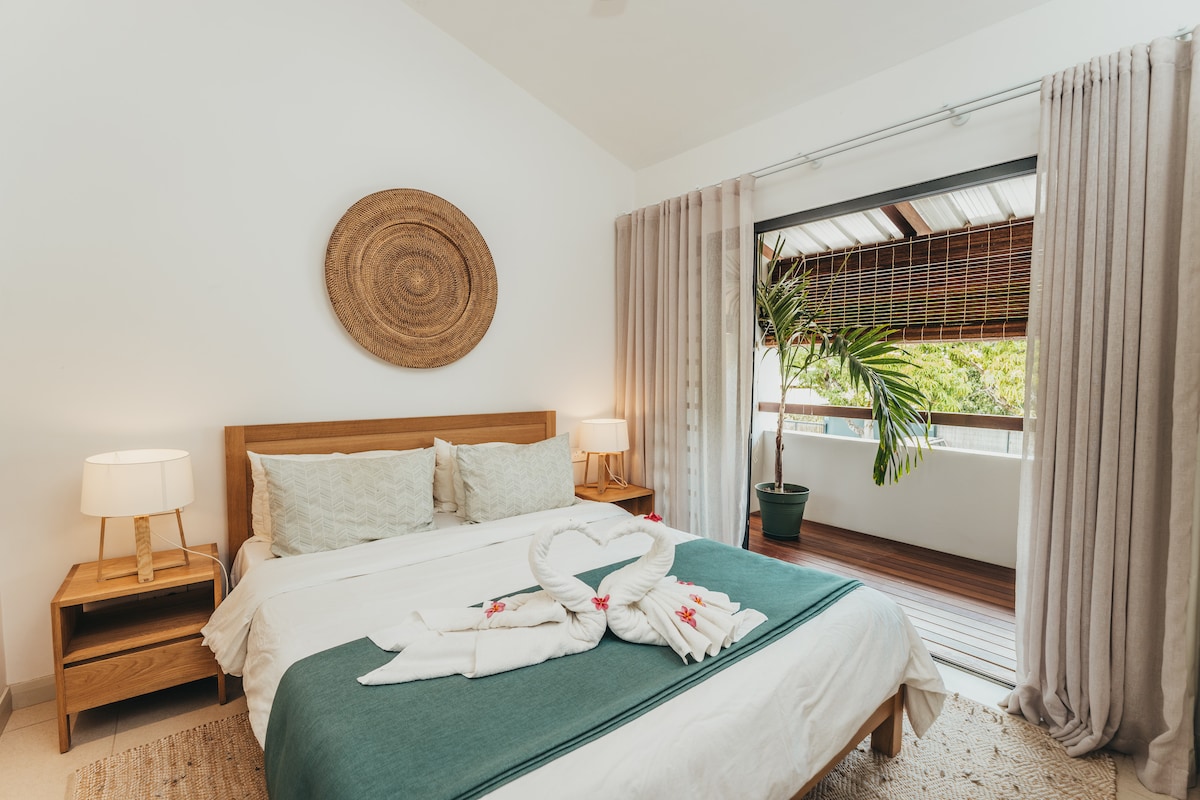 A spacious bedroom is presented with natural light filtering through sheer curtains. The large bed is adorned with a green throw and decorative towels arranged artistically. Nightstands with lamps flank the bed, and a potted plant adds a touch of greenery near the balcony door.