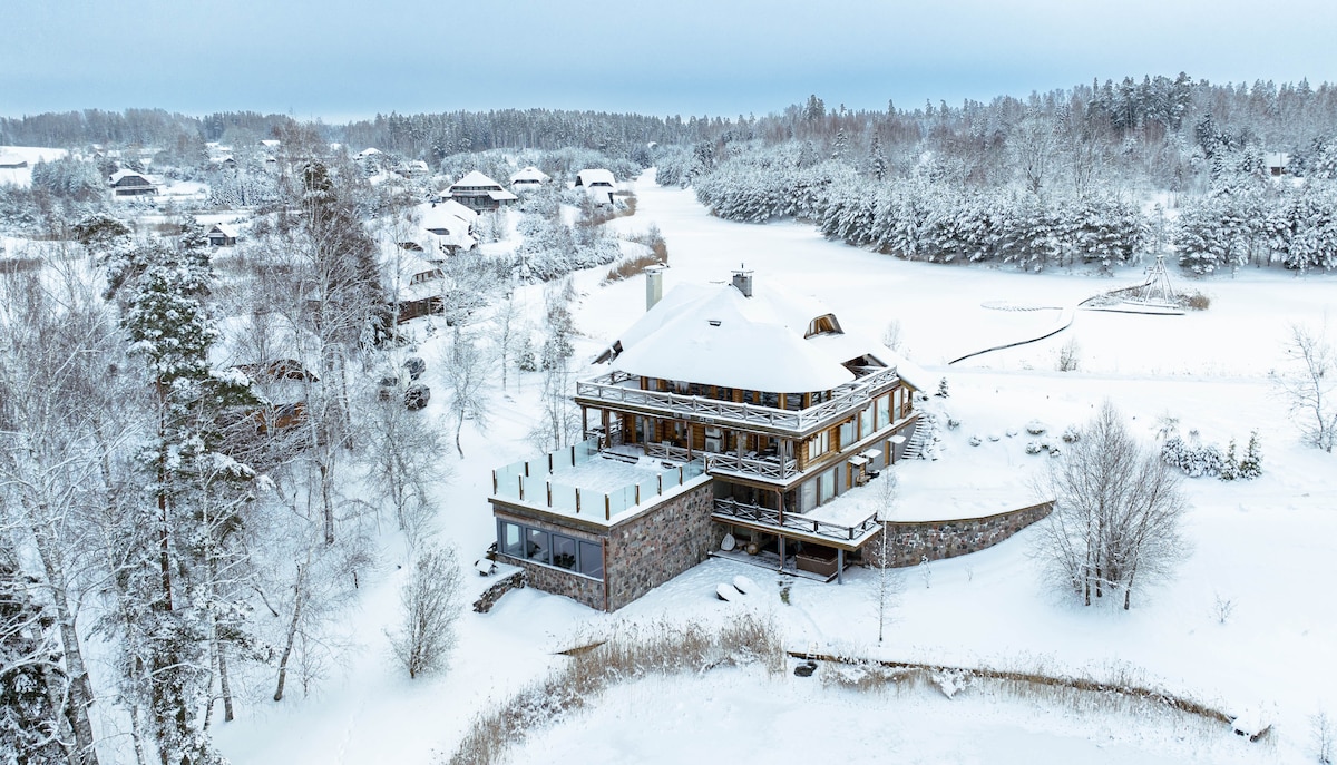 A large wooden structure is surrounded by a snowy landscape, offering a view of nearby trees and a frozen lake. The building features multiple balconies and large windows, suggesting a connection to the natural environment. The serene white snow creates a peaceful backdrop.