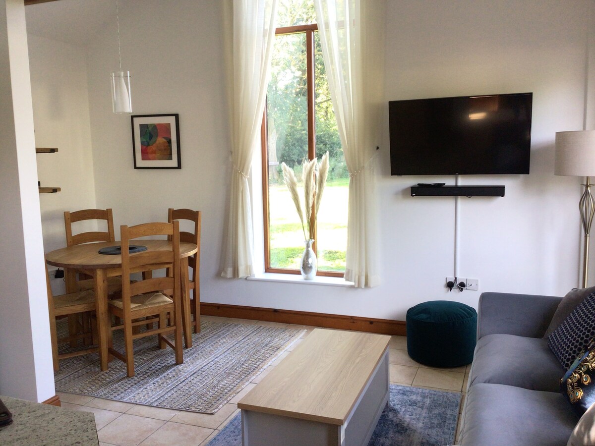 An open-plan living space features a comfortable sofa beside a light wood coffee table. A dining area, consisting of a round table and four chairs, is positioned near a large window, allowing natural light and a view of greenery. A mounted TV is visible on the wall.