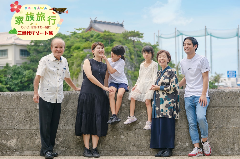 A family of five is standing together at an outdoor location, with a scenic backdrop featuring a castle and greenery. The group includes three adults and two children, with one child seated on a barrier. The adults wear a variety of casual outfits, and the overall mood appears relaxed.