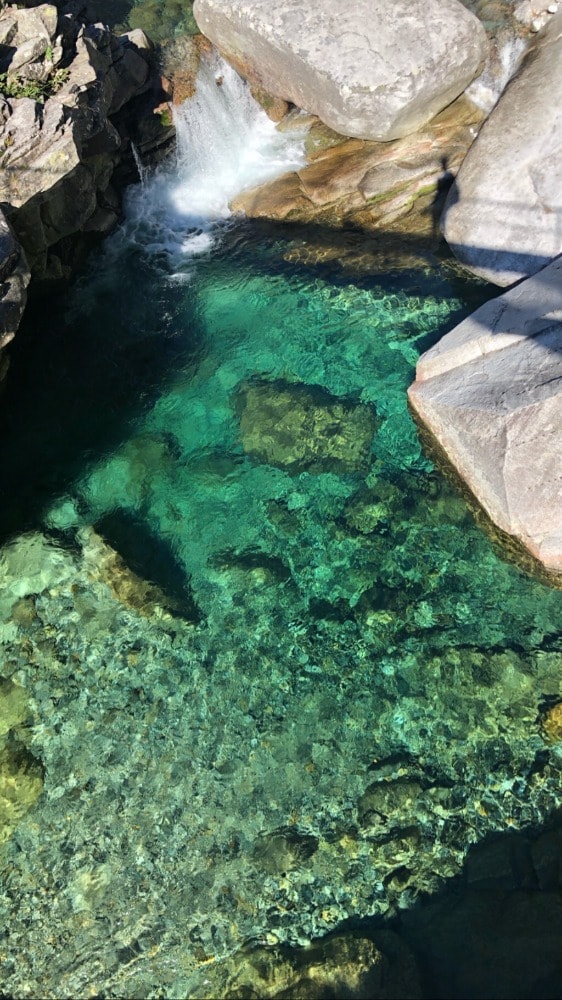 Clear turquoise water flows gently over smooth stones, surrounded by natural rock formations. Sunlight reflects off the surface, creating a serene and inviting atmosphere. The depth of the water varies, revealing colorful pebbles and aquatic plants below.