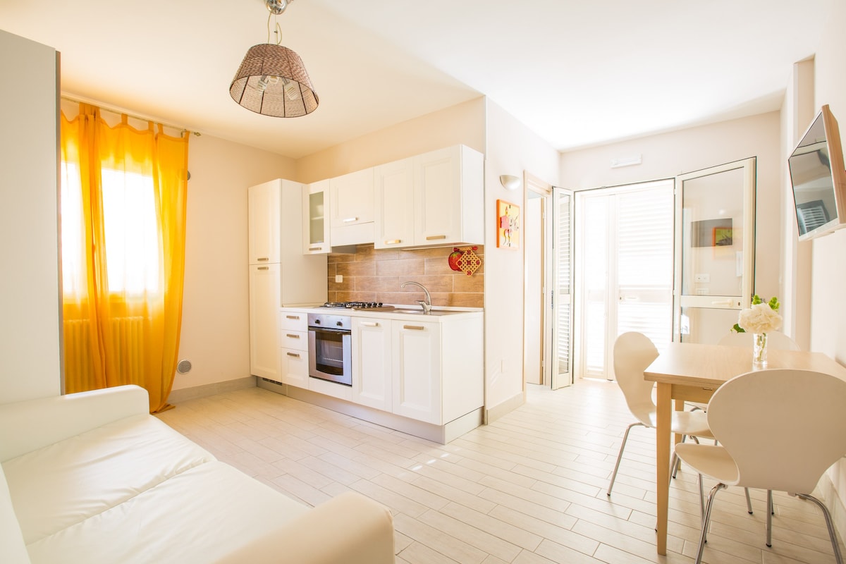 A modern kitchen is visible, equipped with white cabinetry and a stainless steel oven. A light-colored dining table with two chairs is positioned nearby. Natural light fills the space through a doorway leading to an outdoor area, while a vibrant yellow curtain adds a warm touch to the room.