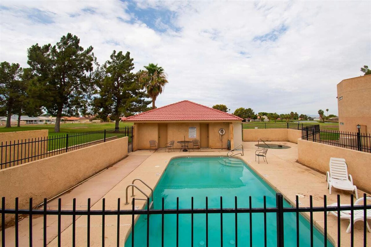 The community pool is surrounded by a low fence, featuring a clear blue water surface and a shallow area. Lounge chairs are placed on the pool deck, with a shaded structure and palm trees visible in the background.