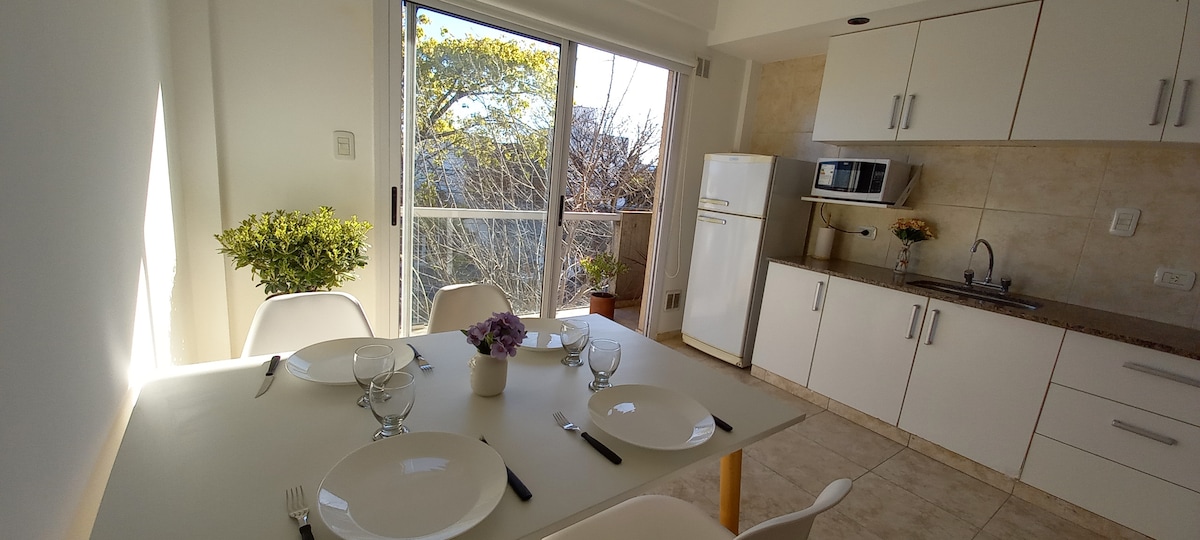 A kitchen dining area features a table set for four, with white plates and utensils. Natural light fills the space through a large glass door that opens to a balcony. A small plant and a vase with flowers add a touch of greenery.