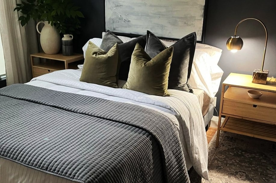 A modern bedroom features a queen bed adorned with plush pillows in shades of green. A cozy grey quilt covers the bed, and a stylish lamp provides warm lighting. Nearby, a wooden nightstand holds decorative items, contributing to the room's contemporary aesthetic.