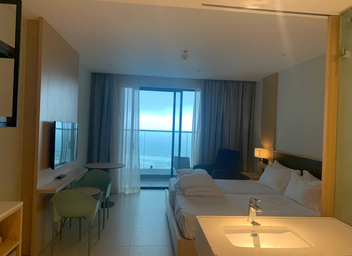 A sleek bedroom features a spacious bed and a small table with green chairs. Large glass sliding doors open to a view of the ocean, allowing natural light to fill the room. A modern television is mounted on the wall opposite the bed.