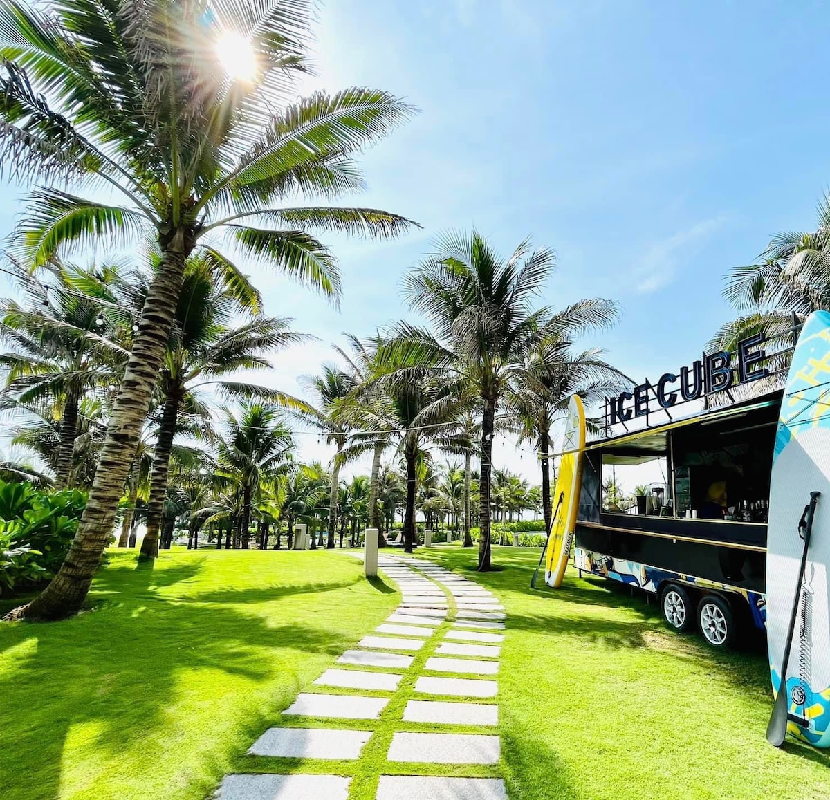 A pathway of stone tiles winds through a lush green landscape, framed by tall palm trees swaying gently in the breeze. A food truck marked 'Ice Cube' sits nearby, accompanied by surfboards leaning against it, inviting relaxation and enjoyment in the outdoors.