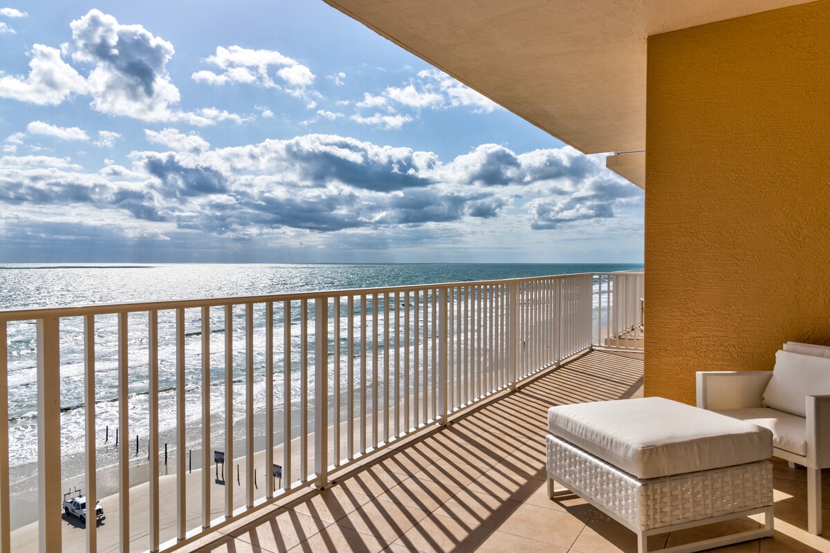A spacious private balcony overlooks the ocean, featuring a comfortable lounge chair and a railing providing unobstructed views of the sandy beach and water. The sunlight casts soft shadows, enhancing the tranquil ambiance of the scene.