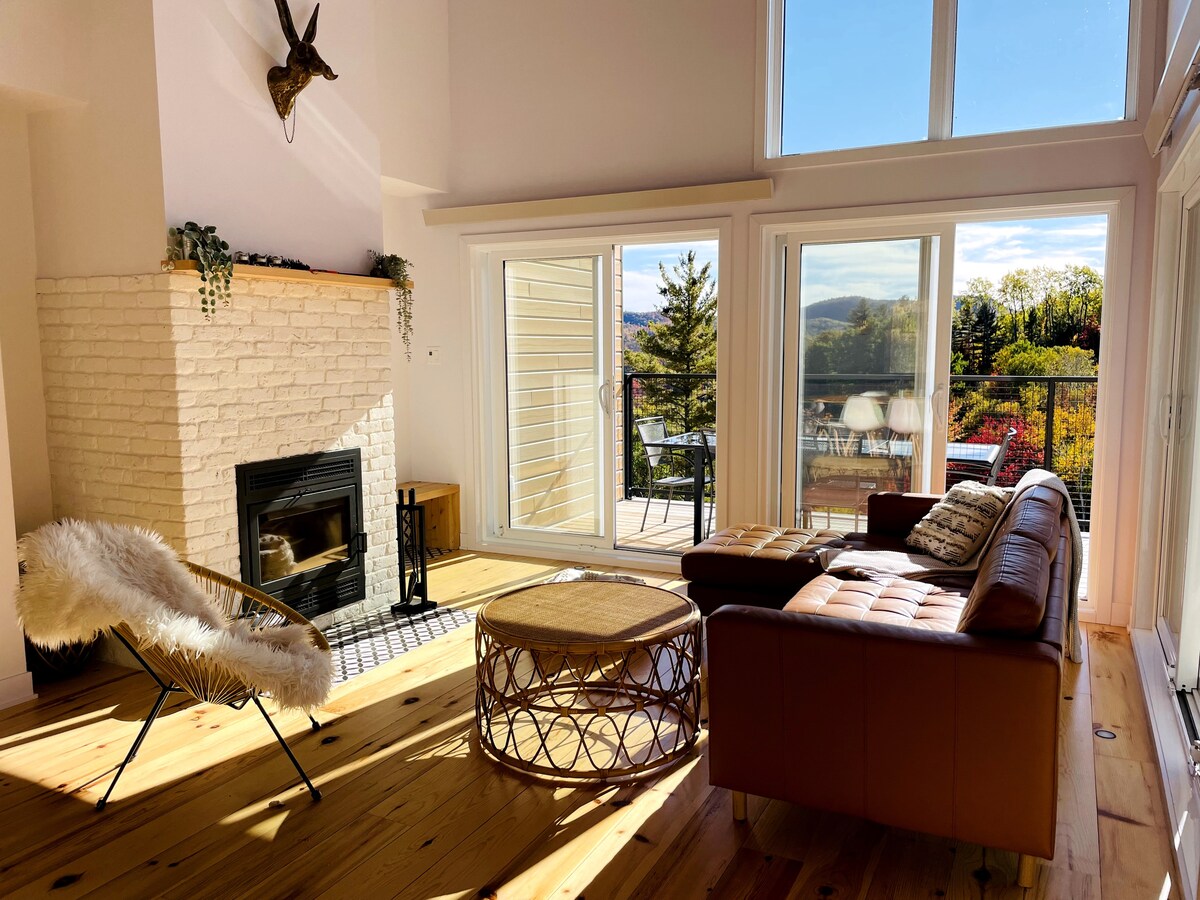 A bright living area features large windows that provide a view of the surrounding landscape. A cozy seating arrangement includes a leather sofa and a round coffee table. A fireplace is positioned against a white brick wall, enhancing the room's warmth.