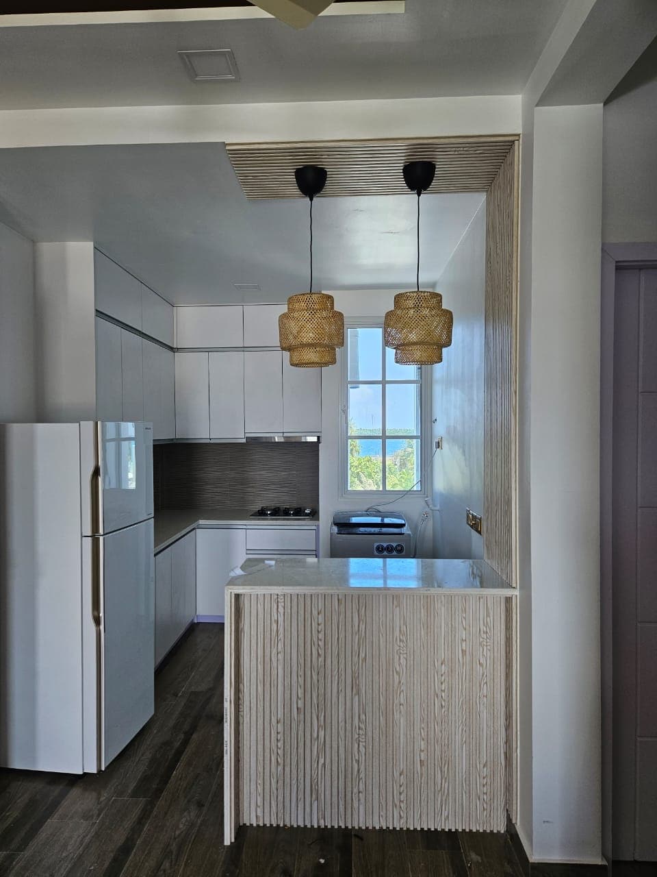A modern kitchen area features sleek white cabinetry and a light-colored countertop. Two pendant lights hang above the counter, providing soft illumination. A large window allows natural light to fill the space, highlighting the clean design of the appliances and cabinetry.