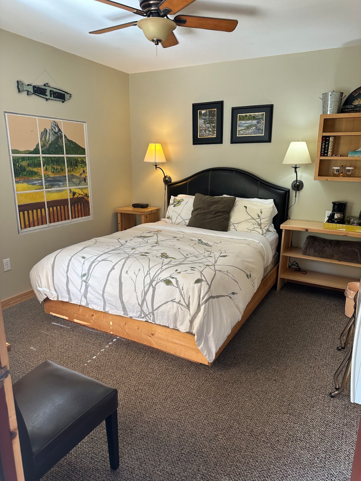 A compact bedroom showcases a wooden bed with a soft white bedding set featuring a nature motif. Two wall-mounted lamps provide lighting beside the bed. A small table and coffee station are positioned against the wall, while a chair sits at the foot of the bed. Nature-themed artwork decorates the walls.