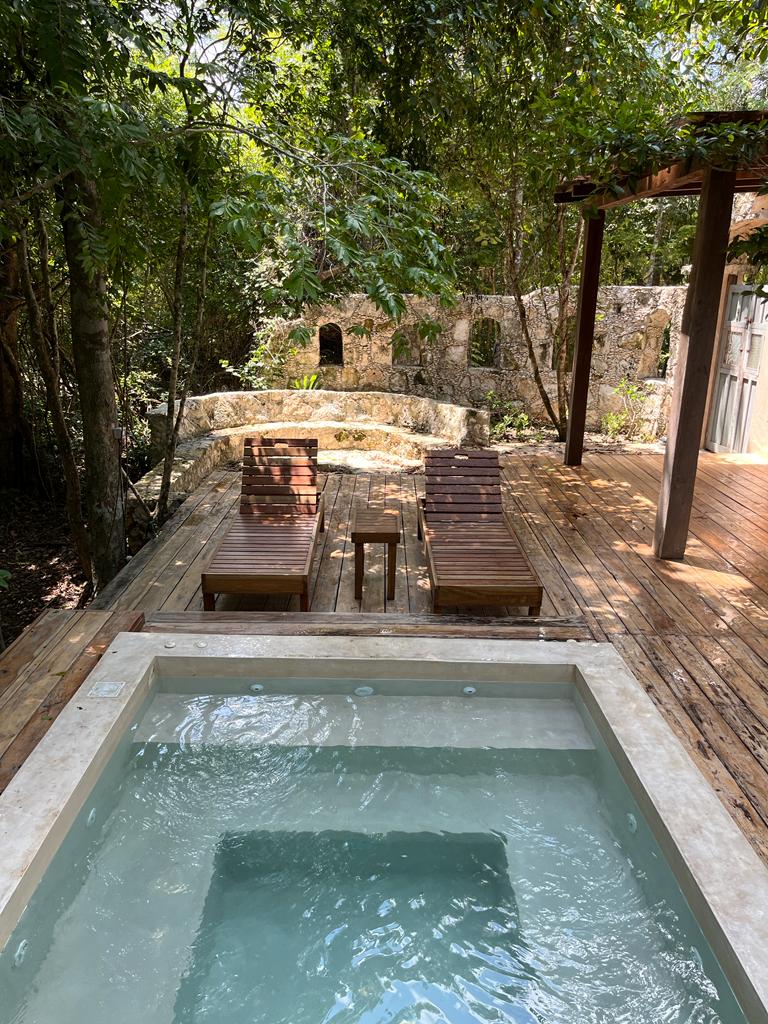 A secluded deck is framed by lush greenery, featuring two wooden lounge chairs beside a small side table. A shallow pool offers a tranquil space, with natural stone walls visible in the background.