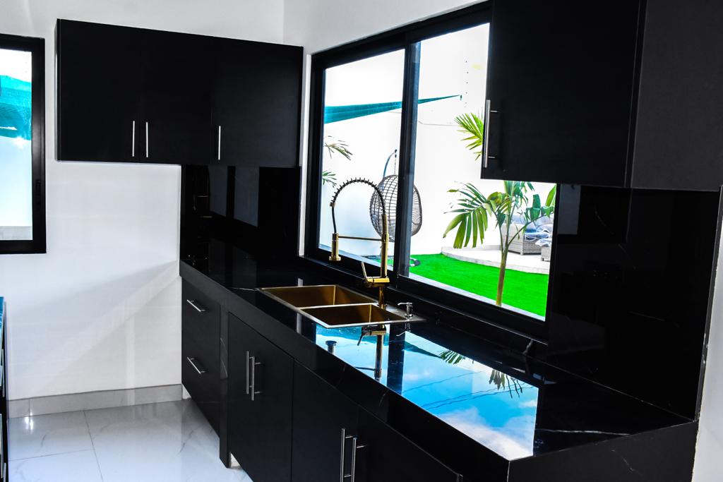 A modern kitchen features sleek black cabinetry and a polished black countertop. A large window allows for natural light, framing views of greenery outside. A stylish sink with a gold faucet is positioned centrally, complemented by minimalist cabinetry above and below.