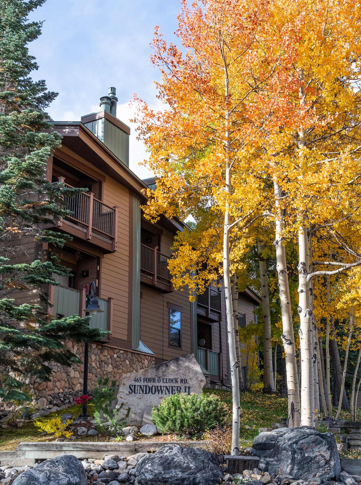 The exterior of a two-level condo is displayed, featuring warm tones and natural stone accents. Vibrant fall foliage frames the scene, with yellow and orange leaves contrasting against the building's neutral colors. A large rock sign identifies the property, while evergreen trees provide a natural backdrop.