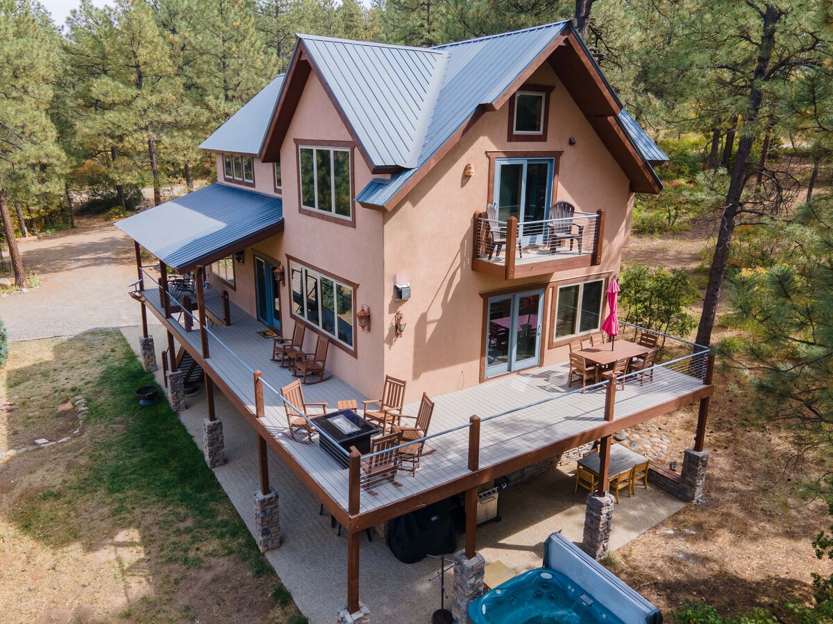The exterior of a large lodge is showcased, featuring a spacious deck with rocking chairs and a hot tub. The building is surrounded by tall pine trees, with a gravel driveway visible nearby. Large windows provide natural light, and a second floor balcony is seen.