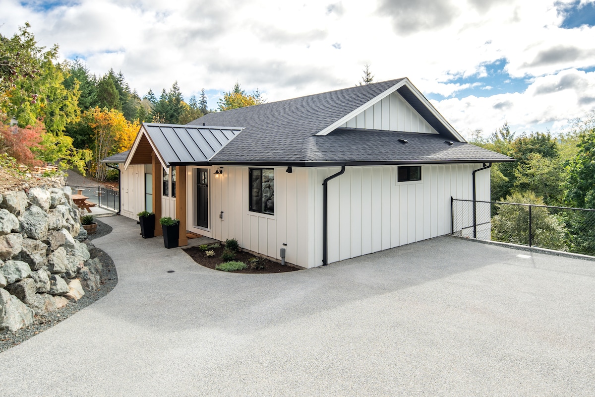 A modern single-level home is showcased, featuring a white exterior with wooden accents. A paved driveway leads to the entrance, bordered by neatly landscaped areas and stone features. Lush greenery and a fence are visible in the background, enhancing the serene environment.