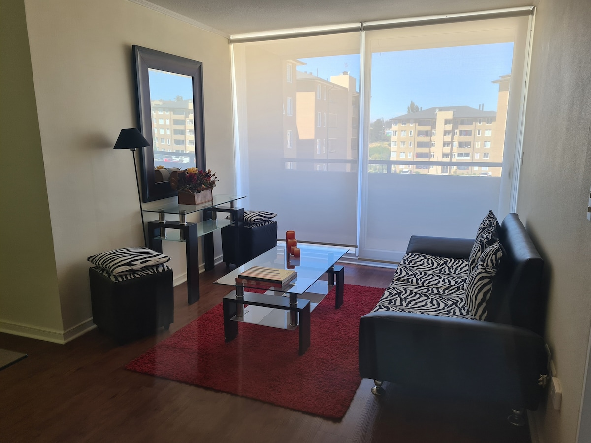 A comfortable living area features a dark sofa adorned with zebra-patterned cushions. A glass coffee table sits on a red rug, surrounded by contemporary stools. Natural light filters through sheer window coverings, creating a bright atmosphere, while a mirror and decorative flowers add a touch of style.