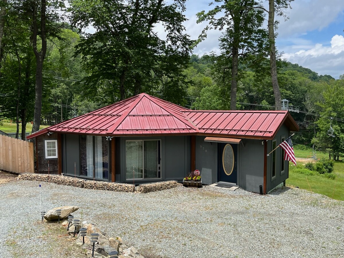 The exterior of a single-story house is shown, featuring a distinctive red metal roof and large windows that reflect the surrounding greenery. A flag is displayed on the front, with a gravel driveway leading to the entrance. Trees and hills are visible in the background.