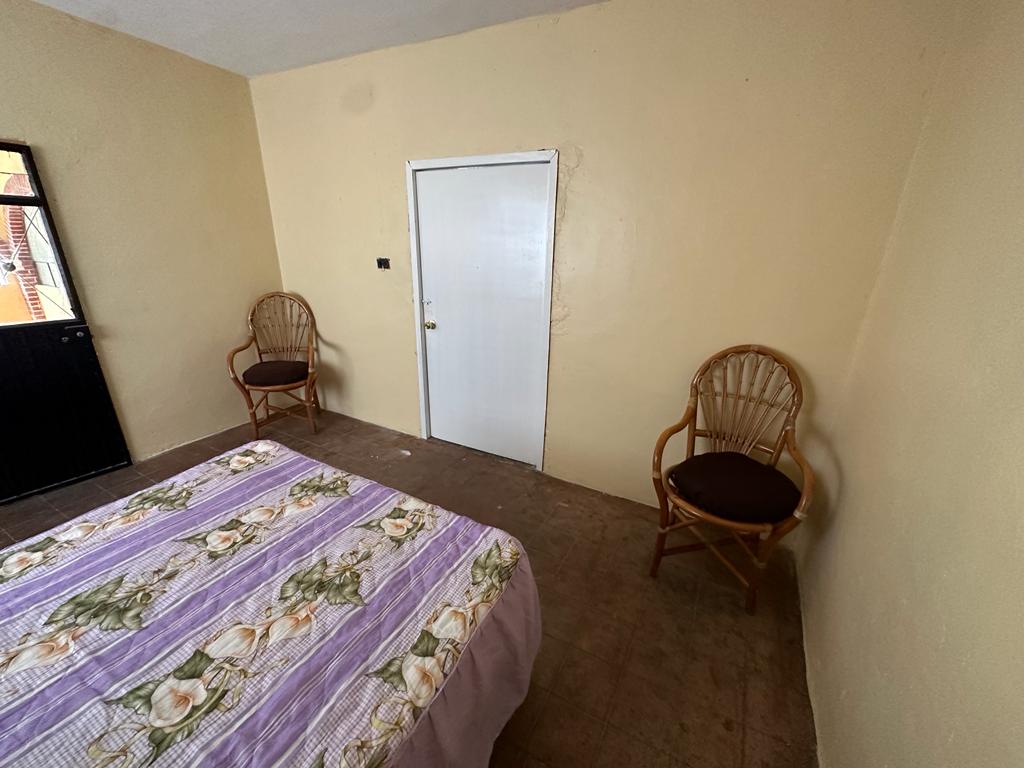 A simple bedroom is presented with light-colored walls and a spacious bed adorned with floral bedding. Two wicker chairs are positioned nearby, offering additional seating. A plain door provides access to another area, and natural light enters through a window to one side.