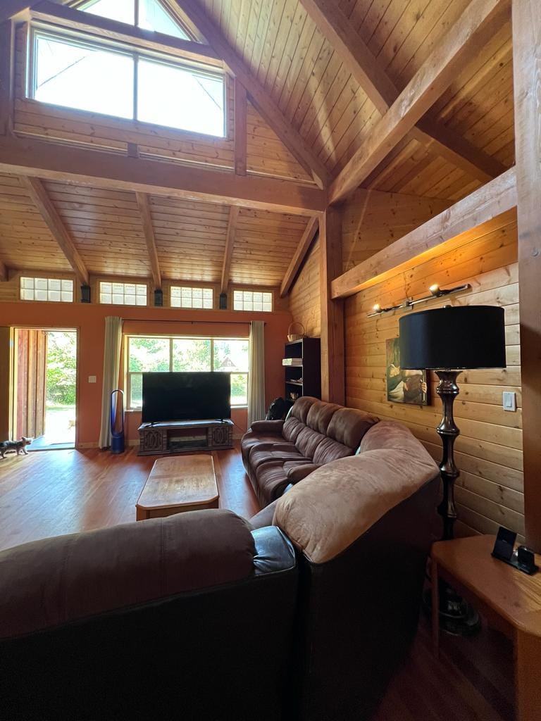 A spacious living area features a high vaulted ceiling with exposed wooden beams. A large brown sofa and a coffee table are arranged, facing a flat-screen TV. Natural light floods the room through tall windows, enhancing the warm wooden decor.