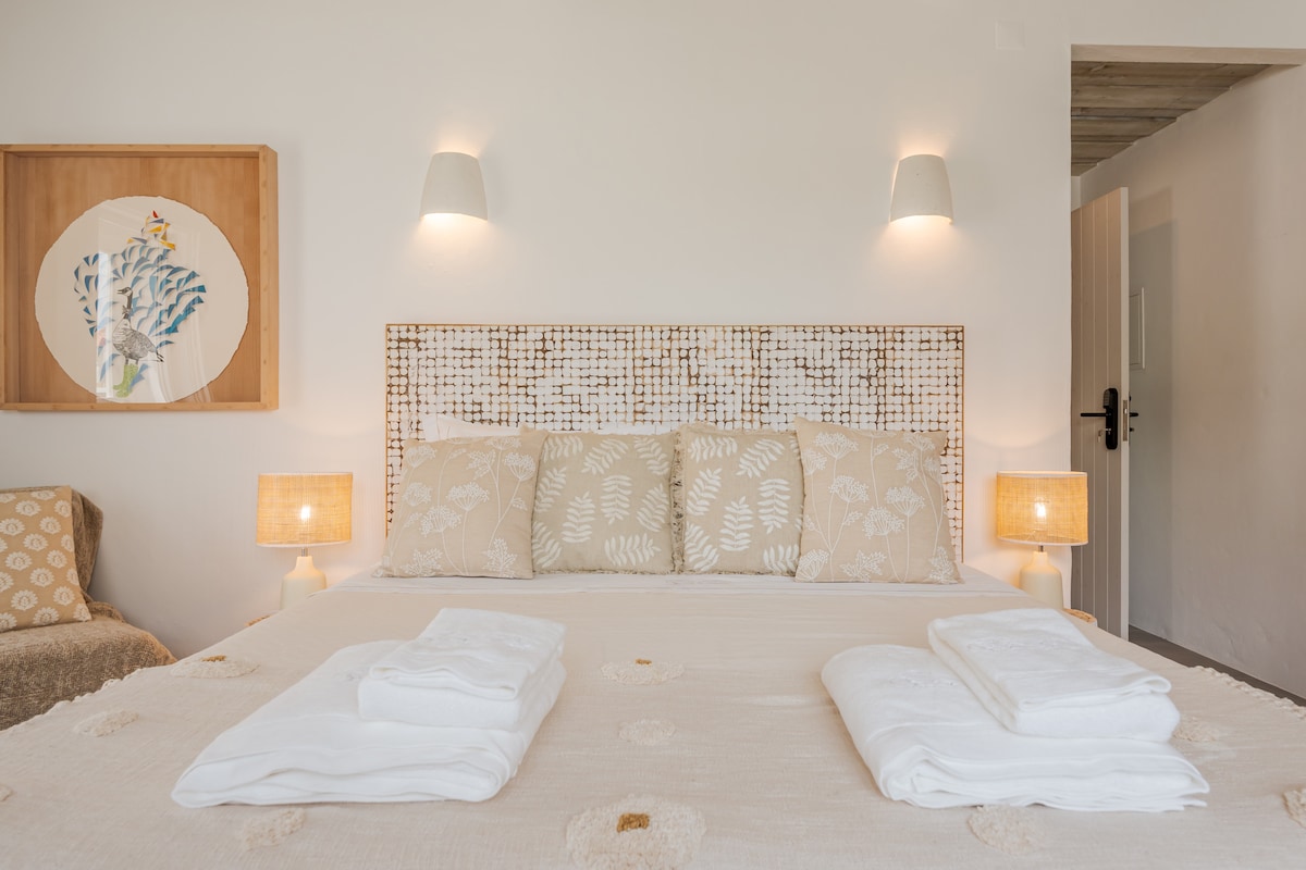 A spacious bed is centered in the room, adorned with soft neutral linens and decorative pillows. Two bedside lamps provide gentle illumination. A piece of wall art featuring natural elements hangs above the headboard, while the overall color scheme evokes a calming and serene environment.