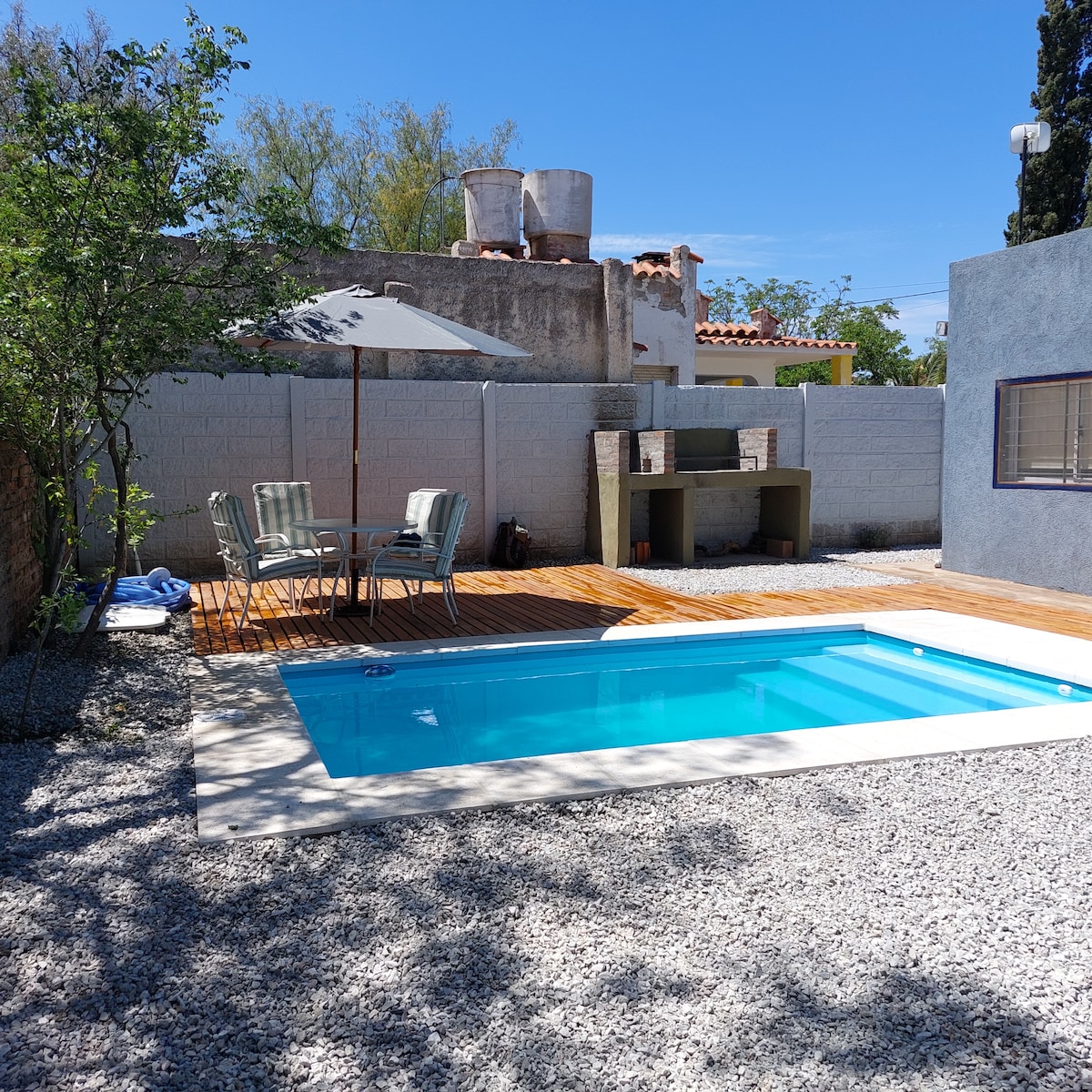 A refreshing outdoor space features a sparkling pool and a wooden deck. A shaded area is provided by an umbrella over a small dining set with four chairs. A grill stands nearby, surrounded by a pebble-strewn surface and greenery.