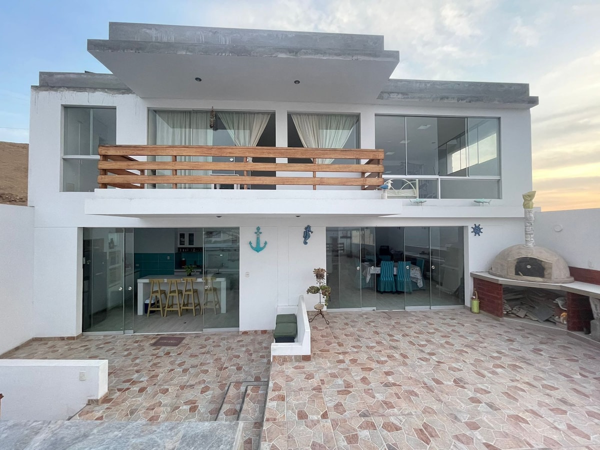 A modern two-story house is presented, featuring large glass sliding doors that open to a spacious patio. A wooden balcony runs along the upper level, while a traditional brick oven is visible on the side. Soft-toned tiles cover the ground, creating a welcoming outdoor space.