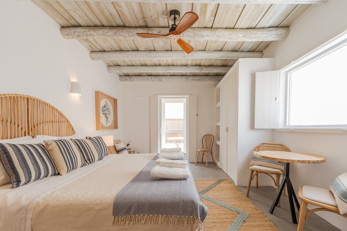 A spacious bedroom features a wooden ceiling with exposed beams, a large bed with decorative pillows, and a soft blanket. A small round table and chair are positioned nearby, while natural light floods in through a bright window, illuminating the warm color palette.