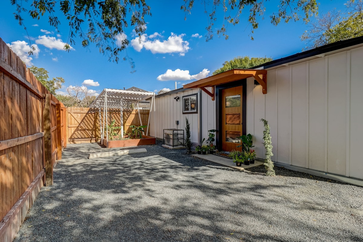 A welcoming entrance to a tidy tiny home is framed by a wooden overhang. The pathway is bordered by gravel, leading to a private garden space with planters. A wooden fence provides seclusion, while blue skies with scattered clouds add to the serene outdoor setting.