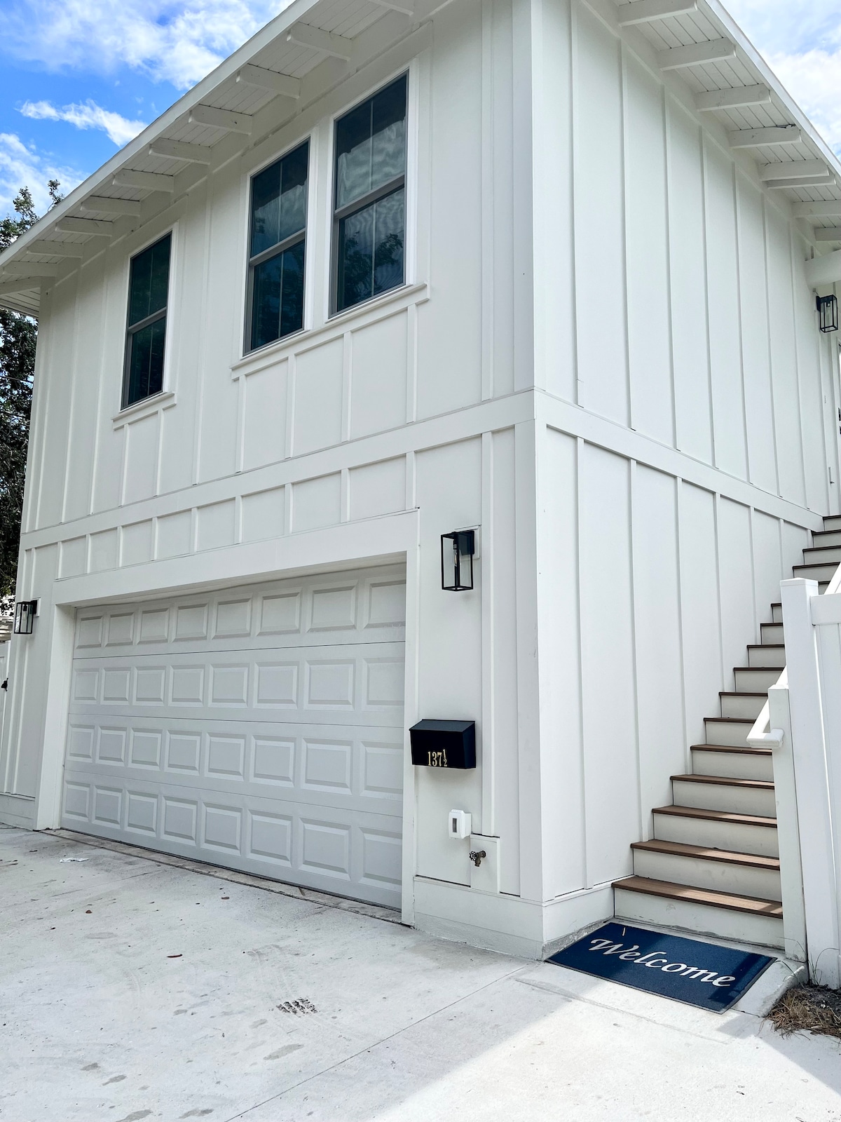 The exterior of the guesthouse is shown, featuring a modern design with white paneling and large windows. A welcoming mat is positioned at the base of the stairway leading to the private entrance above the garage, which has a two-car capacity.