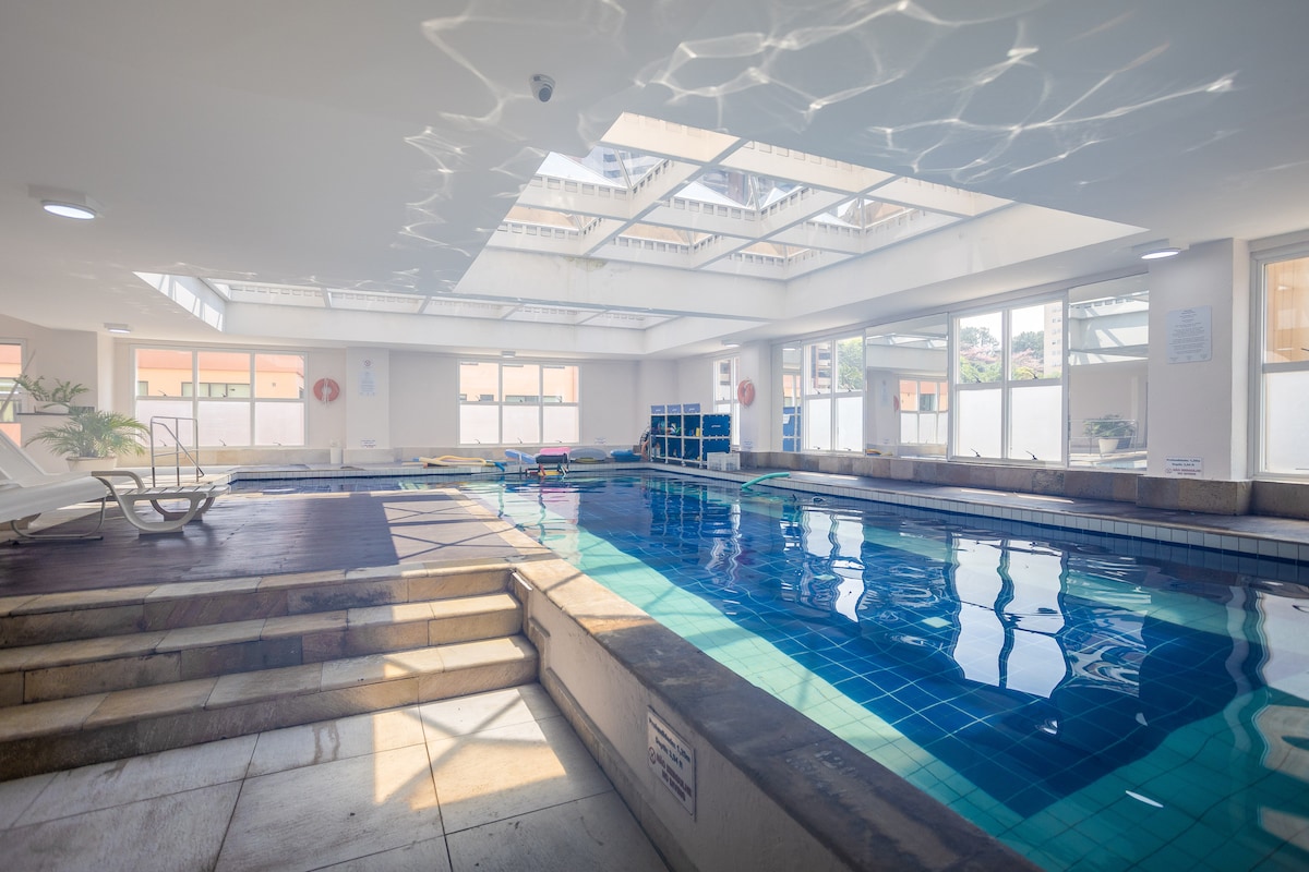 An indoor swimming pool is visible, featuring a partially covered area with natural light filtering through skylights above. Surrounding the pool, lounge chairs are placed for relaxation, and the clear water's surface reflects the overhead light. Safety equipment can also be seen along the walls.