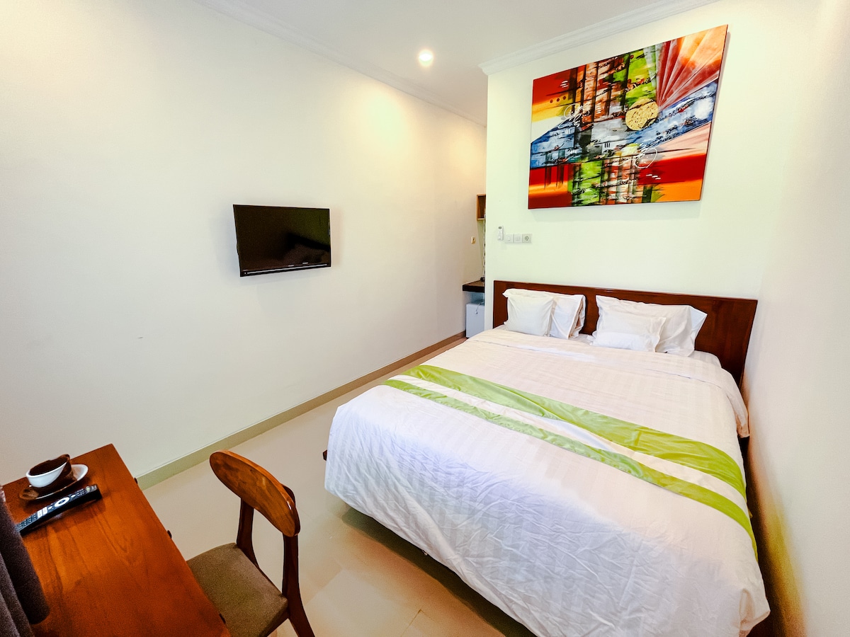 A clean and simple bedroom features a spacious bed topped with white linens and a green accent. A colorful artwork hangs above the bed, while a wall-mounted television is visible. A small wooden desk and chair are positioned near the room's entrance.