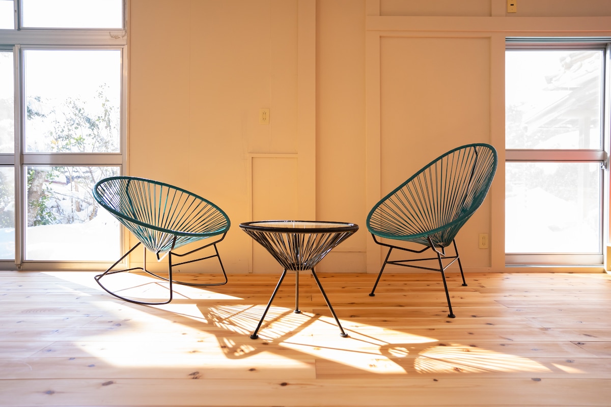 Two modern wicker chairs with teal accents are positioned around a sleek metal table. Sunlight filters through large windows, casting gentle shadows on the wooden floor. The space appears bright and inviting, ideal for relaxation or casual gatherings.